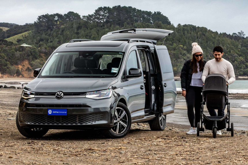 Ein dunkelgrauer Caddy mit geöffneten Türen und Kofferraumklappe am Strand. Daneben ein junges Paar mit Kinderwagen.